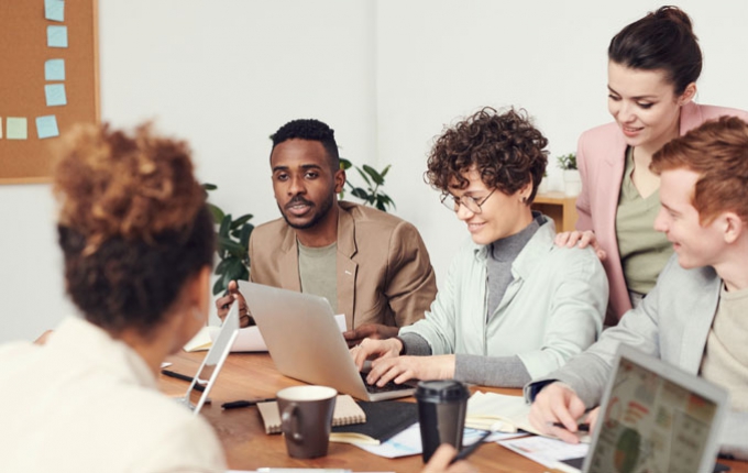 A group of people talk at a table while a woman works on a laptop.