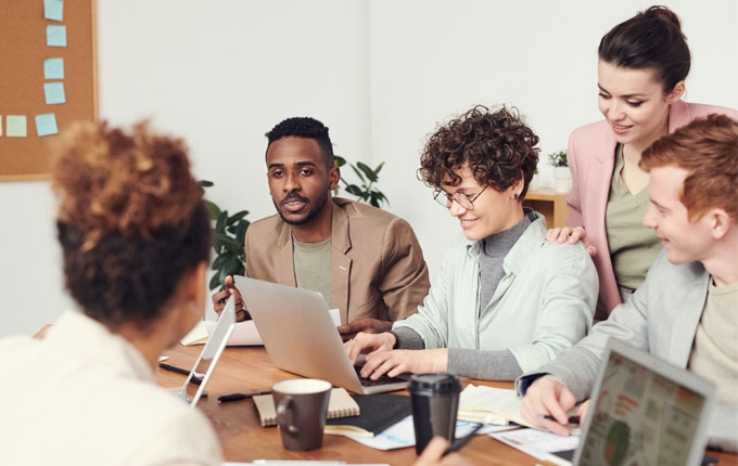 A group of people talk at a table while a woman works on a laptop.